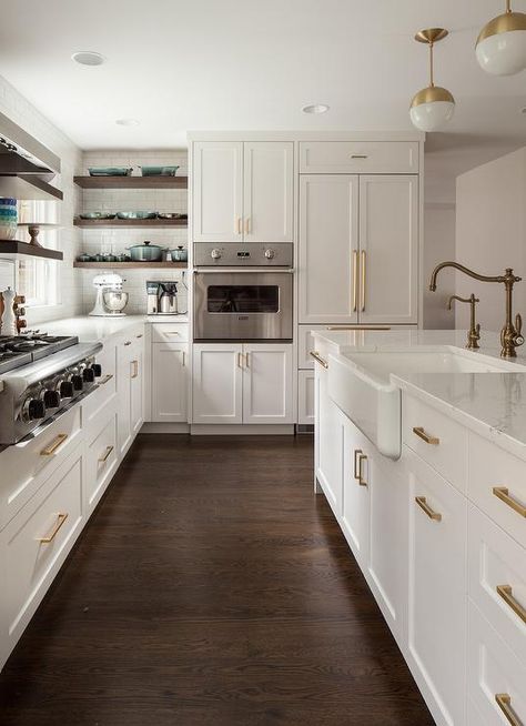 Gorgeous kitchen boasts white and gold pendants illuminating a white center island fitted with a farmhouse sink and antique brass hook and spout faucet. Kitchen With Brown Wood Floors, Off White Kitchen Cabinets With Dark Wood Floors, White Kitchen Dark Brown Floor, White Kitchen Cabinets With Brown Floor, Kitchen Brown Floor White Cabinets, White Kitchen With Dark Brown Floors, Small Kitchen Dark Floor, Dark Floors Kitchen Ideas, Dark Floors With Dark Cabinets