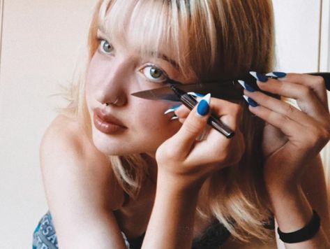 blonde, young girl doing her eyeliner with a knife placed on the side of her eye to help line up the make up. she uses an eyeliner pen and the knife as a ruler against her skin. the photo is edited to look like it was taken off a film camera. Drawing Eyeliner With Knife, Doing Eyeliner With Knife, Knife To Neck Pose, Girl Holding Knife Reference, Woman Holding Knife Reference, Person With Knife Reference, Person With Knife Pose, Person With Knife Pose Reference, Knife To Throat Pose Reference