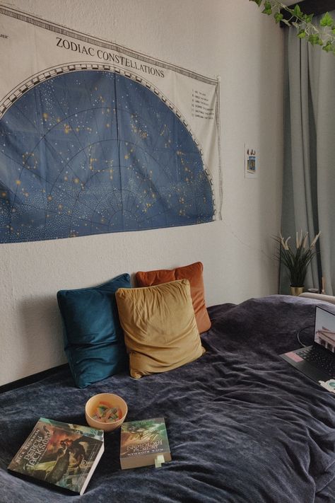 A bed covered with a dark blue velvet blanket, three yellow, blue and orange pillows, the Percy Jackson books, a computer and a yellow bowl with rainbow candy. On the white wall is a tapestry of the zodiac signs and a painting of a parisian street. From the ceiling hangs a vine and the curtains are light blue. Behind the bed is a plant in a golden pot and a red candle light. Astrology Dorm Room, Percy Jackson Themed Bedroom, Aquarius Bedroom Aesthetic, Percy Jackson Room Aesthetic, Percy Jackson Bedroom Ideas, Astrology Themed Bedroom, Percy Jackson Room Ideas, Percy Jackson Decor, Percy Jackson Bedroom