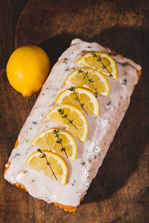 Lemon and Thyme Cake is topped with lemon and thyme glace icing, slices of lemon and sprigs of thyme. It is lying on a dark wood board with a whole lemon to its left. Lemon Thyme Cake, Lemon Thyme Recipes, Thyme Cake, Italian Lemon Cake, Vegan Lemon Cake, Vegetarian Mains, Thyme Recipes, Travel Cake, Dad Birthday Cakes