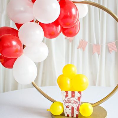 Hula hoop wreath with red and white colored latex balloons and a striped popcorn cardboard box filled with mini yellow latex balloons to form a carnival themed balloon centerpiece. Circus Birthday Party Centerpieces, Circus Centerpieces Diy, Circus Theme Party Centerpieces, Carnival Party Centerpieces Diy, The Greatest Showman Centerpieces, Carnival Theme Centerpieces Diy, Carnival Birthday Centerpieces, Elegant Circus Theme Party, Dumbo Birthday Party Decorations