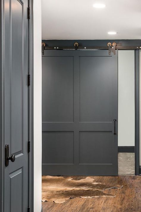 A charcoal gray barn door on rails opens to a basement boasting a brown cowhide rug. Raccoon Fur Paint Color, Dark Grey Baseboards, Raccoon Fur Benjamin Moore, Benjamin Moore Gunmetal, Wolf Gray Benjamin Moore, Benjamin Moore Raccoon Fur, Transitional Basement, Grey Painted Furniture, Basement Door