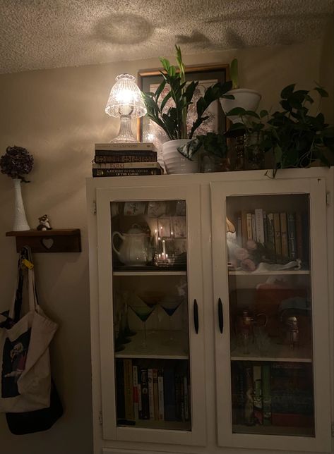 entryway with china cabinet full of thrifted glassware and books witj small crystal lamp and plants on top China Cabinet Styling Ideas, Cabinet For Glassware, Thrifted Glassware, Top Of Cabinets, Dining Room Updates, China Hutch, China Cabinet Display, Room Update, Crystal Lamp