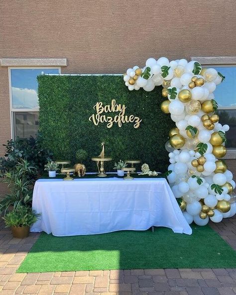 Welcome your baby with a garden inspired outdoor table setup to elevate your baby shower party décor. Use a white tablecloth for your tabletop and arrange gold cake and cupcake stands over it to place your sweet confectioneries. Maximize its beauty by using faux plants that match your green table runner. Opt for an artificial grass wall for your backdrop adorning it with uniquely set white and gold balloons. Add finishing touches with some large sized fake plants that’ll exude refreshing vibes. Cake Table Backdrop Birthday, Green And White Decorations Party, Expo Backdrop, White And Gold Balloons, Garden Party Decorations Diy, Greenery Party, Party Decorations Table, Artificial Grass Wall, Balloons Cake