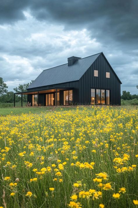 Modern barndominium charcoal black facade in stunning farmhouse meadow with yellow flowers. Check out the coolest dark barndominium houses that combine rustic charm with modern sophistication, enhancing a living space with bold hues and sleek designs. Contemporary Barndominium Ideas, Barndominium With Screened In Porch, Black And Cedar Barndominium, Barndominium Lake House, Barndominium 2 Story, Barndominium Landscaping, Steel Homes Metal Houses, Dark Barndominium, Dark Barndominium Exterior