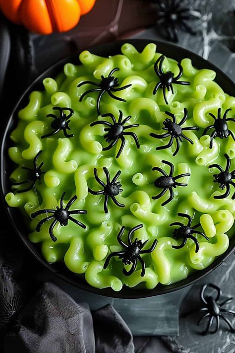 Close-up of a bowl filled with vibrant green mac and cheese, garnished with black plastic spiders. The background includes Halloween-themed elements like pumpkins and cobwebs, adding to the festive atmosphere. Mac And Cheese For Kids, Fun Halloween Drinks, Halloween Pasta, Spooky Halloween Desserts, Spooky Dinner, Halloween Lunch, Halloween Themed Food, Pumpkin Ale, Spooky Food