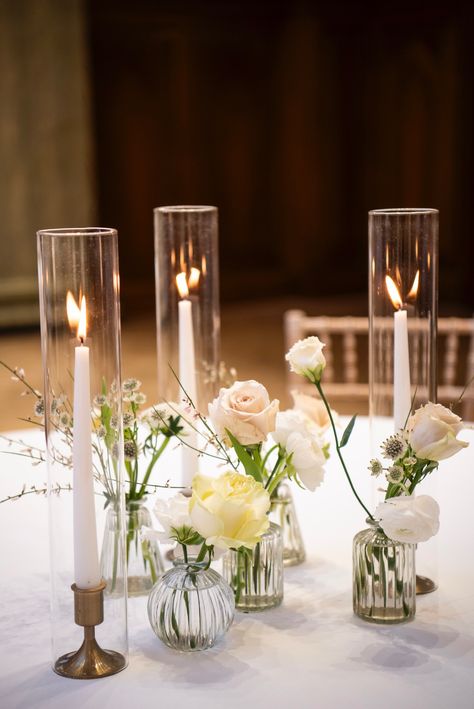 Three taper candles in hurricane covers with pretty flowers in glass bud vases for a wedding table centrepiece. Tapered Candles In Glass Cylinders, Candles And Bud Vases, Floral Wedding Arrangements, Table Centrepiece Wedding, Wedding Table Decorations Candles, Wedding Reception Candles, Farm Table Wedding, Christmas Wedding Table, Bud Vases Wedding