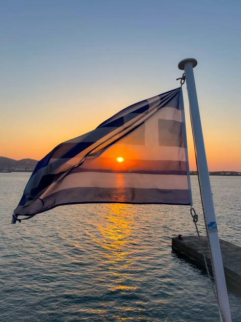 Multiple colors of bright orange red and yellow sunset of the ocean of Greece with the Greek flag shaded over flying in the wind Greece Flag Aesthetic, Greek Flag Aesthetic, Greek Sunset, Ef Ultimate Break, Greece Sunset, Aesthetic Greece, Trip Fashion, Greece Flag, Summer Ball