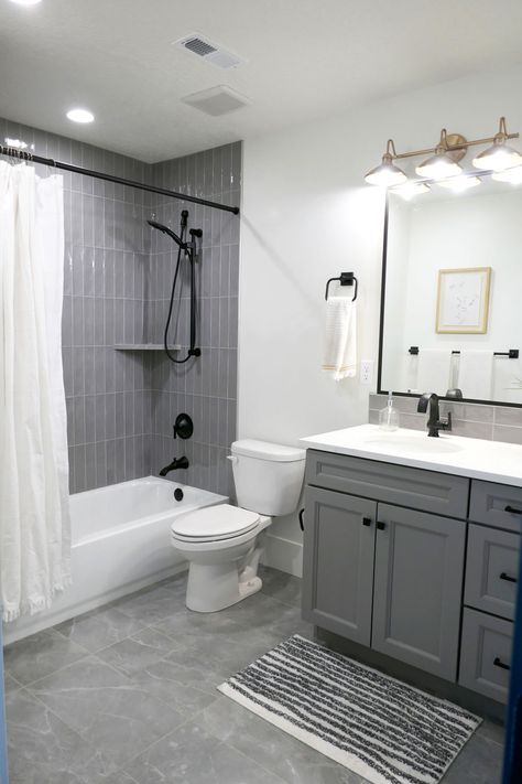 A bathroom with a tub/shower combo that has tub to ceiling gray glass subway tile installed vertically. The toilet is between the tub and single-sink vanity, which has darker gray cabinets and white Quartz countertops. The floor has large rectangular tiles that are gray with white veins. White Bathroom With Grey Floor, Modern Bathroom Grey And White, Gray Floor White Walls Bathroom, White And Dark Grey Bathroom, Gray Toilet Bathroom, Grey Tile Shower Bathroom, Gray Sink Bathroom, Small Bathroom White And Grey, Small Bathroom With Grey Vanity