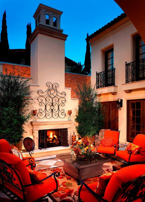 Iron armchairs with vibrant red cushions enclose a cozy sitting area and accent the iron railings on the stucco home exterior. A large outdoor fireplace serves as the focal point in front of a large wicker coffee table, creating an inviting conversation area. Mediterranean Patio, Living Pool, Outdoor Sitting Area, Mediterranean Style Homes, Casas Coloniales, Outdoor Fireplaces, Mediterranean Decor, Spanish Style Homes, Diy Outdoor Decor