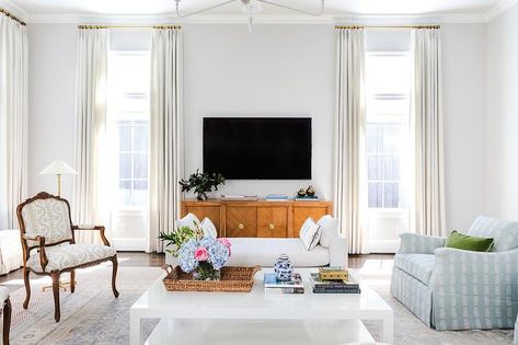 White curtains hang from brass curtain rods in front of windows flanking wooden TV console located beneath a wall mount TV. Tv Between Windows, Blue Ottoman Coffee Table, Windows Design Ideas, Brown Wooden Bed, White Settee, Windows Design, Blue And White Pillows, Tv Wall Decor, Cottage Living Rooms