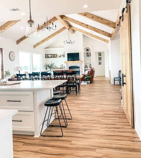 A large open concept kitchen, living, and dining room is filled with natural light from the wall of windows. A vaulted ceiling with wood beams runs the length of the space with two modern, black metal chandeliers hanging from the center beam. Over the kitchen island hangs a glass lantern pendant light. Open Concept Vaulted Ceiling, Open Concept Kitchen Dining Living Room, Vaulted Ceiling Ideas, Vaulted Ceiling Lighting, Beams Living Room, Vaulted Ceiling Kitchen, Open Concept Kitchen Living Room, Vaulted Ceiling Living Room, Open Kitchen And Living Room