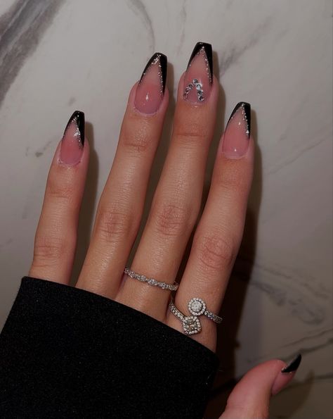 The picture is showing acrylic nails. There is a hand in front of a modern background. The nails are black French tips with silver glitter around the edges of the French tip. The middle finger has gems on it. Black Nails With Glitter Design, Black French Tip Nails With White Line, Modern Almond Nail Designs, Nails French Ideas Black, Black French Tip Nails With Silver, Black Glitter French Tip Nails Coffin, Black French Tip Acrylic Nails With Gems, Black French Acrylic Nails Coffin, Edge French Tip Nails