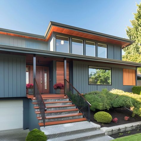 This mid-century modern split-level house boasts a stunning blue-gray vertical siding with warm wood accents. Large upper-level windows fill the interior with natural light. The inviting entryway features wide stairs leading to a covered porch with a wooden door, flanked by potted plants. Meticulously maintained landscaping includes trimmed bushes and vibrant flowers, enhancing the home's curb appeal and creating a welcoming atmosphere. Modern Tri Level Exterior, Split Level Paint Exterior, Split Entry Remodel Exterior, Split Level Home Exterior, Wide Stairs, Split Level Remodel Exterior, Split Entry Remodel, Mid Century Renovation, Bush House