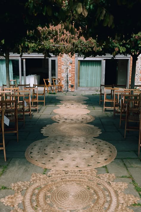 This shows an image showing a ceremony area at an outdoor wedding. On the aisle are different jute rugs lined to accentuate the aisle. Alongside the aisle are vintage folding chairs. Isle Rugs Wedding, Rugs Isle Wedding, Jute Rug Aisle Runner Wedding, Rugs For Outdoor Wedding, Round Rug Wedding Ceremony, Rugs At Wedding Ceremony, Outdoor Wedding Rugs, Rugs Aisle Wedding, Rugs For Wedding Aisle