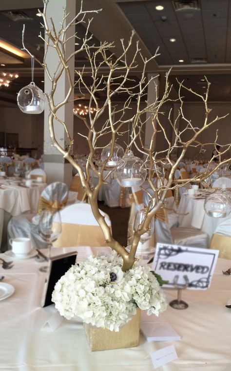 Reception centerpieces. Arrangement includes gold sprayed manzanita branches, hydrangeas, gold square vase and hanging globes. Check out www.luminousbloomsartistry.com for more ideas. Cream And Gold Centerpieces, Gold Tree Centerpiece, White And Gold Banquet Decor, Elegant Birthday Centerpieces, Gold White Centerpieces, Derby Centerpieces, White And Gold Centerpieces, Gold Tree Branch Centerpiece, Winter Wedding Table Decorations