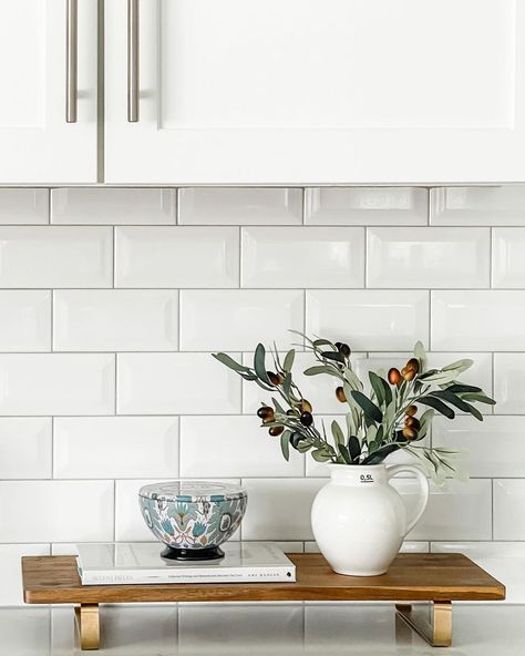 A white kitchen cabinet hangs over a white subway tile backsplash with light grey grout. Green branches in a white pitcher sit on a wooden tray next to a blue bowl on a white book. The tray is placed on the white countertop. Kitchen Counter Styling Ideas, Kitchen Wall Covering, Counter Styling, White Kitchen Counters, Kitchen Counter Styling, Modern Kitchen Counters, Gray Grout, White Countertop, Light Gray Cabinets