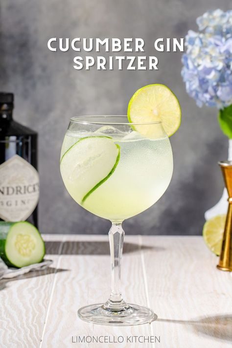 Side view of a Cucumber Gin Spritzer cocktail on a white wood countertop. The drink is in a tall stemmed glass with a round bowl. It is light green and semi translucent, and the glass has a strip of cucumber inside and a lime wheel on the rim. In the background are bar tools and hydrangea flowers along with ingredients. Fancy Drink Garnish, Cucumber Spritzer Vodka, Cucumber Drink Alcohol, Cucumber Spritzer, Gin Cocktails Summer, Light Summer Cocktails, Cucumber Gin Cocktail, Drink Garnish, Gin Drink Recipes