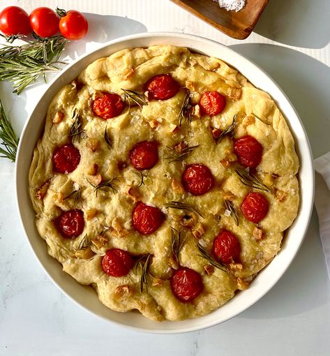 There is a large round white serving dish with baked flat bread in it. There are cherry tomatoes, rosemary leaves, pieces of roasted garlic, and salt flakes on to of the top of the bread. There is 3 cherry tomatoes and 2 sprigs of rosemary laying behind the serving dish. Items are on a white marbled surface. Rosemary Recipes, Bread Art, Salt Flakes, Focaccia Bread, Flat Bread, Rosemary Leaves, No Knead, Best Side Dishes, Dried Tomatoes