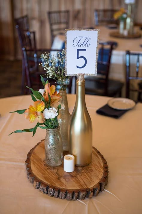 Wedding reception centrepiece on wood round with gold wine bottle for table number holder Wine Bottle And Wood Centerpieces, Table Number Wine Bottle, Wine Bottle Table Decorations, Wine Bottle Centrepiece, Gold Bottle Centerpiece, Wine Bottle Table Decor, Centerpieces With Wine Bottles, Gold Wine Bottle Centerpiece, Wine Bottle Centerpieces For Wedding