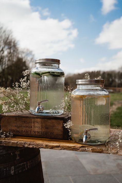 This image shows a drinks station at a wedding. The focus is on two glass dispenser containers, with cucumber and lemon water in them. One of the containers is displayed on a crate, with dried flowers surrounding the bar. Double Drink Dispenser, Drink Dispensers Wedding, Drink Dispenser Stand Diy, Drink Display Wedding, Wedding Coolers, Bakery Theme, Drink Dispenser Stand, Diy Pedestal, Drinks Dispenser