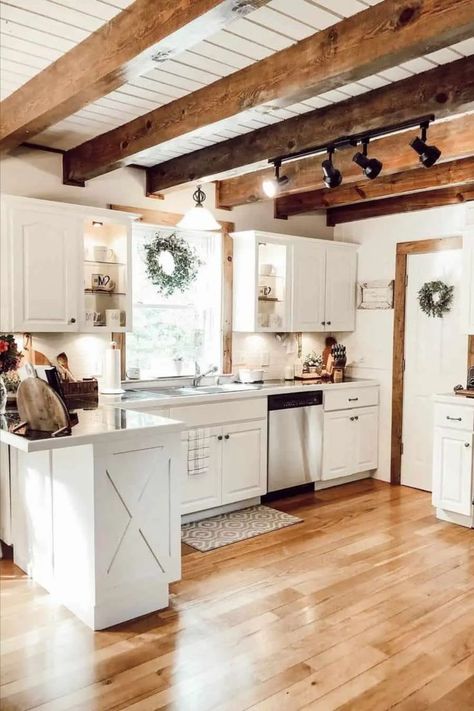 This modern kitchen showcases innovation with its pristine white countertops and suspended shelves, creating a striking contrast with the polished wooden flooring. Delicate decorative wreaths add a touch of charm, and the wooden ceiling beams, suspended beneath the painted-white ceiling, house adjustable black metal lights, creating an atmosphere of pure elegance. Beams Living Room, Farmhouse Kitchen Decor Ideas, Rustic Farmhouse Kitchen Decor, Modern Farmhouse Ideas, Wooden Beams Ceiling, Farmhouse Kitchen Design, Rustic Farmhouse Kitchen, Farmhouse Kitchens, Kitchen Ceiling