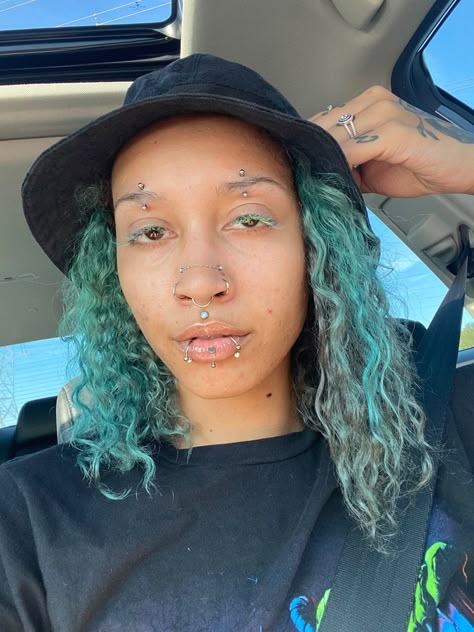 Picture of a person with curly hair displaying their piercings. They have an silver curved barbell in the center of each eyebrow. They have double opal high nostril piercings, each sitting above a nostril ring. They have a septum that hangs in the center of they’re nose just above an opal stud in the dip of the top lip. They also have a labret in the center of they’re bottom lip framed by a ring on each side of the bottom lip. Snakebites And Labret, Double Vertical Eyebrow Piercing, Eyebrow Piercing Double, Labret And Snakebites, Centered Eyebrow Piercing, Snakebites And Vertical Labret, Double Brow Piercing, Vertical Labret And Snakebites, Hoop Eyebrow Piercing