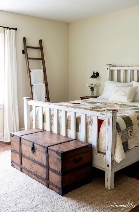 Featuring a distressed white wooden bedframe, this room is the epitome of farmhouse charm. Pale cream walls complement a hand-quilted patchwork bedspread. A vintage ladder serves as a towel rack, adding rustic flair. A large wooden trunk at the foot of the bed doubles as a storage bench. Cozy Bedroom Design, Patchwork Bedspread, Wooden Trunk, Cozy Bedrooms, Vintage Ladder, Country Bedding, Wooden Trunks, Quilted Patchwork, Cream Walls