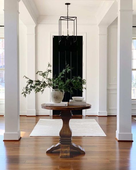 This elegant foyer with white columns is lit by a black lantern pendant light and boasts a black front door. A stained wood pedestal round entryway table is placed in front of the door. A large glass vase filled with greenery accessorizes the top of the table. Round Entryway Table, Entryway Round Table, Line Interior Design, Round Entryway, Wood Pedestal Table, Round Foyer Table, Round Entry Table, Entryway Table Ideas, Gallery Wall Staircase