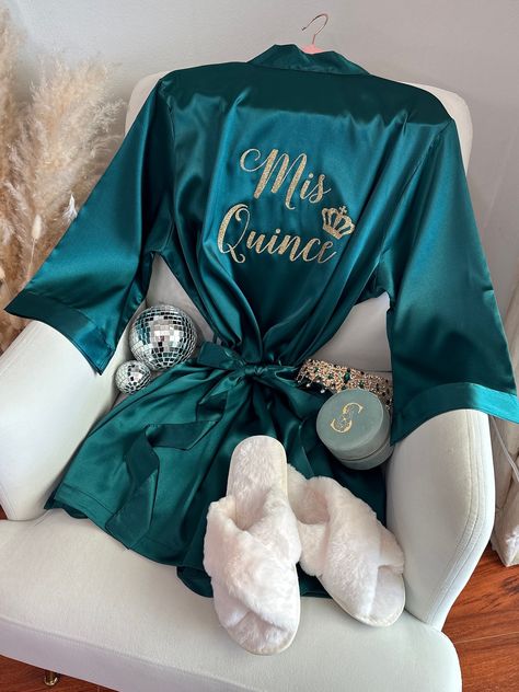 "Celebrate your special Quinceañera with our beautiful Mis Quince Satin Robe, a perfect addition to your memorable day! Crafted from high-quality soft silky satin, our Quinceañera robe ensures a luxurious and comfortable experience while getting ready for the celebration. Our robes come with inner side pockets!  Customize your robe with glitter text or mirror metallic for that extra touch of glamour! Choose from a variety of glitter or metallic options to make your Mis Quince Robe uniquely yours Emerald Green Quinceanera Surprise Dance Outfit, Dark Teal Quinceanera Theme, Emerald Green Quince Robe, Dark Green Chambelanes Outfits, Quince Robe Ideas, Mis Quince Robe, Dark Green Quince Decorations, Teal And Gold Quinceanera Theme, Quinceañera Gift Ideas