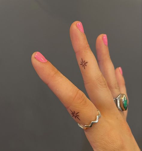 Tiny starburst finger tattoo. Two tattoos on middle finger and pointer finger. Gray wall background. Cute Side Finger Tattoos, Small Tattoos On Fingers Simple, Small Tattoos Finger Women, Dainty Finger Tats, Star Tattoos Finger, Sparkle Tattoo On Finger, Tiny Starburst Tattoo, Stars Tattoo On Finger, Starburst Hand Tattoo