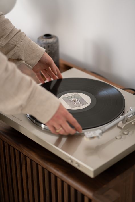 A person wearing a beige long-sleeve shirt is placing a vinyl record onto a turntable. The turntable, which is on a wooden cabinet, has a silver tonearm and modern design. In the background, a decorative ceramic vase is visible. The scene is softly lit, creating a warm and cozy atmosphere. Stereo Stand, Audio Cabinet, Record Player Table, Record Player Stand, Vinyl Player, Turn Table Vinyl, Vinyl Storage, Functional Storage, Record Player