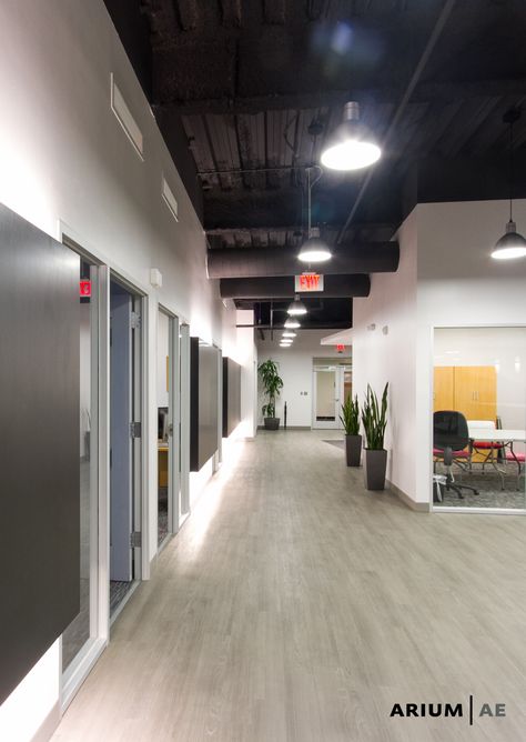 Corridor in an office space, with black laminate accent wall with light coves, exposed ceiling painted black, vinyl wood-look laminate floors Black Ceiling Office Design, Office Exposed Ceiling Design, Exposed Office Ceiling, Black Ceiling Office Interiors, Office With Black Ceiling, Exposed Ceiling Office Design, Black Office Ceiling, Dark Office Ceiling, Dark Ceiling Office