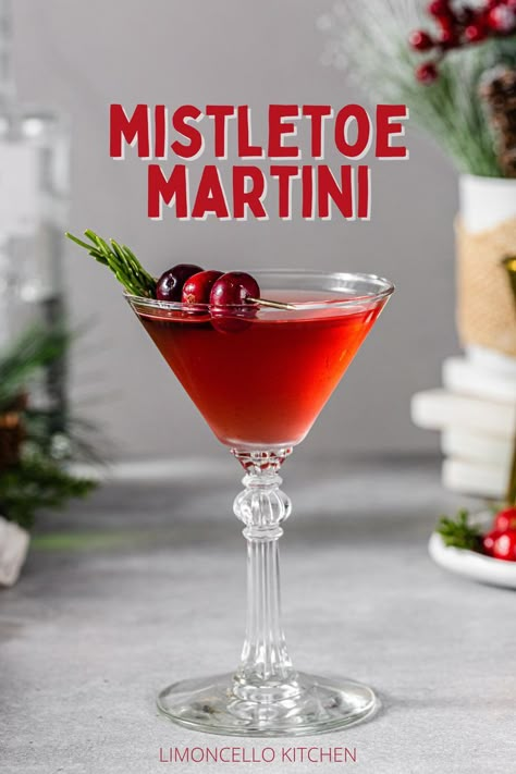 Side view of a Mistletoe Martini cocktail in a martini style glass. The red colored cocktail is garnished with three cranberries on a gold cocktail pick and a sprig of fresh rosemary. To the right of the glass is a plate of cranberries and rosemary along with a gold jigger. On the left are evergreen sprigs, a bottle of gin and a green bottle of vermouth. Text above the drink says “Mistletoe Martini” in red capital letters. Below the drink it reads “Limoncello Kitchen” in red capital letters. Christmas Martinis Holiday Drinks, Christmas Martini Holiday Drinks, Mistletoe Martini Recipe, Christmas Gin Drinks Cocktail Recipes, Pomogranette Martini, Mistletoe Martini, Rosemary Garnish, Tito’s Christmas Cocktail, Blueberry Martini