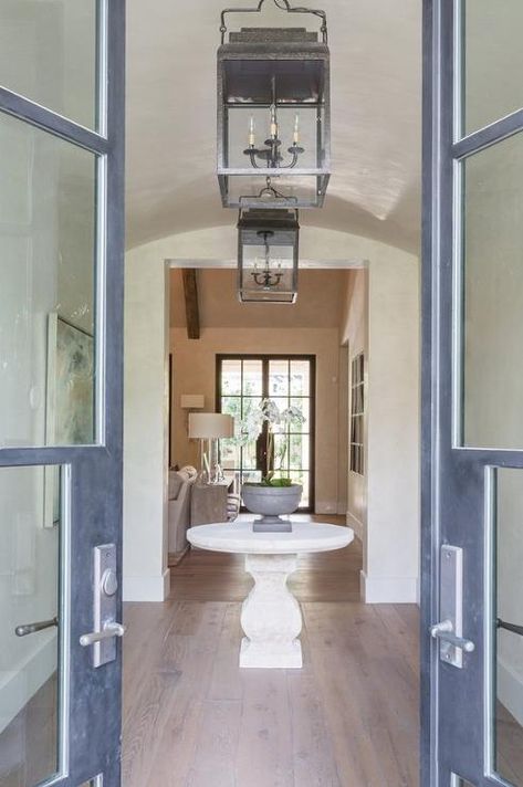 Glass double doors open to a foyer featuring a round stone pedestal table placed in the center of the room illuminated by a vintage lantern. Symmetrical Entryway, Foyer Round Table, Round Stone Table, Glass Double Doors, Stone Pedestal, Foyer Stairs, Double Door Entryway, Round Glass Table, Front Entryway