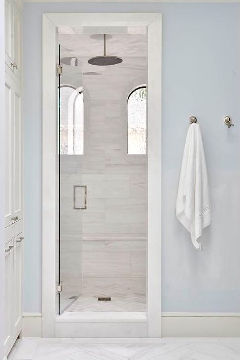 Light blue wall paint surrounds a seamless glass door opening to a walk-in shower fitted with marble herringbone floor tiles mounted beneath a round rain shower head. Herringbone Floor Tiles, Pale Blue Bathroom, Large Bathroom Design, Marble Herringbone Floor, Jenkins Interiors, Marble Herringbone Tile, Dream Condo, Blue Bathroom Walls, Herringbone Tiles