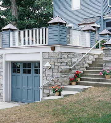 Integrate with Landscape  The homeowners of this house took advantage of their garage's location behind their house and added a deck above it that connects to the house. The stone exterior of the structure blends well with the rest of the landscaping. The blue door matches the trim and siding found on the house further integrating the design Patio On Top Of Garage, Porch On Top Of Garage, Under Deck Garage, Balcony Over Garage Ideas, Deck Over Driveway, Flat Roof Garage Ideas, Tandem Garage Ideas, Deck Over Garage, Lower Level Garage