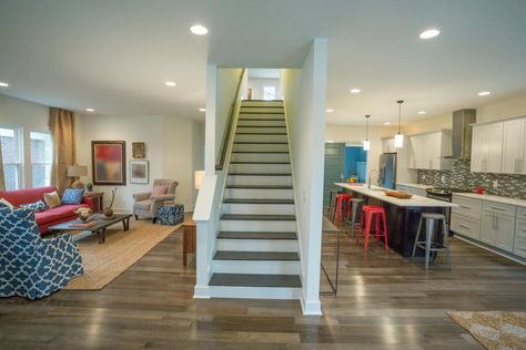 Recessed lighting illuminates this open concept living space with all-over brown hardwood floors and a center staircase. Red and blue upholstered furniture decorates the living room along with a sisal area rug, while red metal stools and a gray mosaic tile backsplash complete the otherwise white kitchen. Recessed Lighting Ideas Living Room, Open Stairs In Living Room, Center Staircase, Recessed Lighting Ideas, Kitchen Lighting Over Island, Remodeled Kitchens, Recessed Lighting Living Room, House Renos, Open Concept Kitchen Living Room