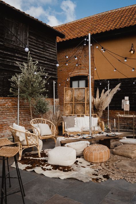 This image shows an outdoor barn wedding lounge area. The area consists of cane sofa, cane chairs, floor cushions, low level tables, cane bar stools, vintage screen. On the floor are cow hide rugs and jute rugs. Surrounding the lounge area are potted live trees and dried flowers. Above the area are string lights. Outdoor Wedding Props, Wedding Vignette Ideas, Casual Wedding Seating, Outdoor Wedding Furniture, Cowhide Rug Wedding, Wedding Ceremony Seating Ideas, Lounge Area Wedding, Cowhide Wedding, Wedding Lounge Seating