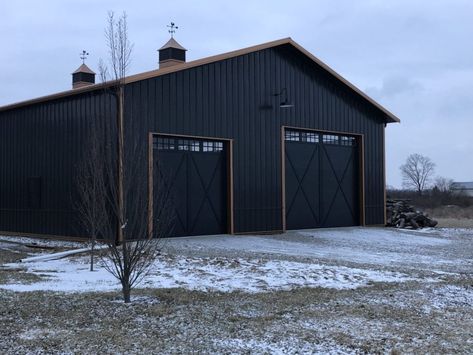 This 50' by 72' machine storage and garage was built with black steel panels and a bronze trim. The bronze roof and dual cupolas with weather vanes on top of the building add a distinguished final look. 80x100 Metal Building, Black Outbuilding, Black Morton Building, Black Steel Roof House, Black Metal Building With Wood Accents, Black And Copper Metal Building, Black Workshop Exterior, Black Metal Shop Building, Black Steel Building