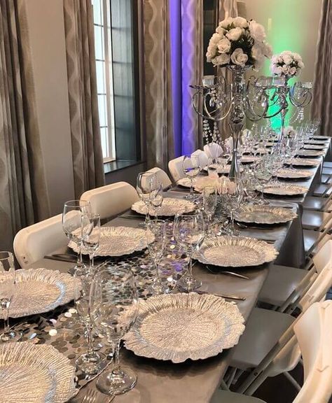 Set up a lavish reception table to impart grandeur & luxury to your wedding festivity. Drape the table with a pretty silver tablecloth & spread a silver big sequin Payette runner on top for a charismatic allure. Arrange white roses on top of silver candelabras with crystal hanging drops to ooze a lovely floral flair. Also, use silver reef chargers and embellish the chairs with white cushions to finish off your majestic silver themed table decor. White And Silver Table Decor, Silver Table Decor, Silver Wedding Decor, Silver Table Setting, Ceremony Space Decor, Anniversary Dinner Ideas, Silver Wedding Reception, Silver Tablecloth, Silver Wedding Decorations