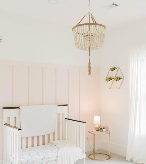 A white and pale pink nursery includes a board and batten wall behind a crib accessorized with blankets and a patterned mattress. A beaded chandelier hangs above the room that includes a gold metal side table and wall art. Pink Board And Batten Wall, Pink Board And Batten, Sherwin Williams Intimate White, Intimate White, Blush Pink Paint, Pink Painted Walls, Nursery Paint Colors, Grown Up Bedroom, Sherwin Williams White