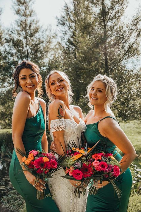 Bridesmaids in emerald green one shoulder dresses with bride in strapless lace boho chic wedding gown holding bright colourful bouquets Emerald Green Bridesmaid Dresses With Colorful Flowers, Bridesmaids Bouquets With Green Dresses, Emerald And Pink Bridesmaid Dresses, Emerald Green Wedding With Bright Flowers, Bouquet With Emerald Green Dress, Dark Teal Summer Wedding, Ocean Wedding Bridesmaid Dresses, Festival Wedding Bridesmaid Dresses, Flowers For A Green Dress