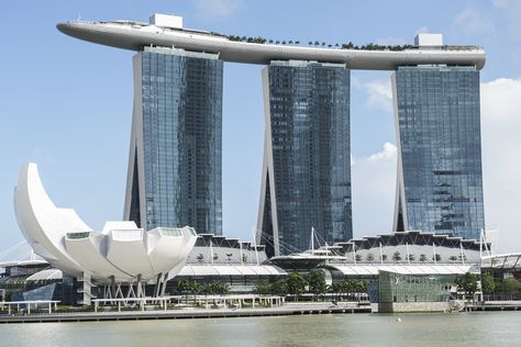 The monumental Marina Bay Sands resort was completed in 2011 on the Singapore waterfront. The centerpiece of the complex is the hotel, which is made up of three 55-story towers connected by the Sands SkyPark. The cantilevered space includes a swimming pool, gardens, a public observation deck, restaurants, and jogging paths. The complex also has a casino, convention center, theaters, and the Museum of ArtScience. Famous Architecture Buildings, Moshe Safdie, Singapore Architecture, World Famous Buildings, Sands Hotel, Famous Architecture, Building Photography, Famous Buildings, Perfect Itinerary