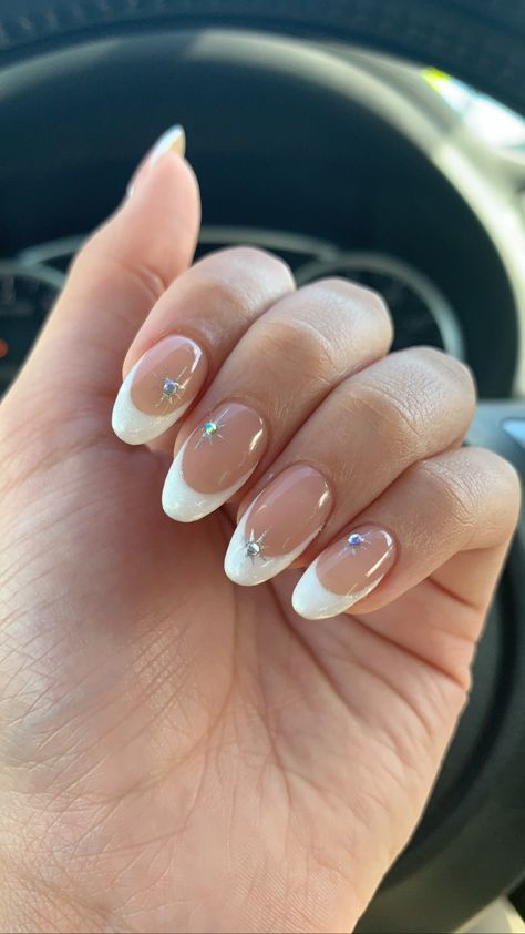 Almond shaped nails with a white glitter color. Chrome twinkle design with translucent gems in the center of the twinkles. White And Gem Nails, French Tip Gel Nails With Rhinestone, French Mani With Gems, French Tip With Jewels Nails, Nails Confirmation, Oval Nails With Gems, Chrome Nails With Diamonds, Chrome Nails Gems, French Nails With Crystals