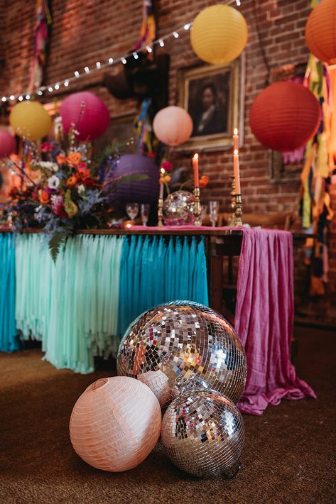 This image shows a table at a barn wedding. The table is decorated with colourful streamers on the side. On the floor beside the table are large disco balls and round paper lanterns. On the table are brass candlesticks, disco balls and colourful flower arrangements. Above the table are round, colourful paper lanterns and a string of lights. Disco And Flowers, Wedding Table Decor Disco Balls, Disco Ball At Wedding Reception, Colorful Wedding Decor, Disco Garden Party, Disco Ball Wedding Decor, Wildflower Disco Ball Wedding, Wedding Tent Disco Ball, Disco Ball Colorful Wedding