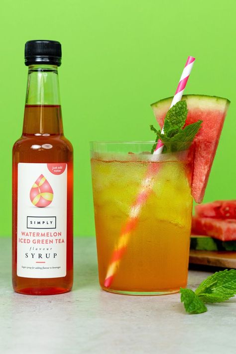 A glass of watermelon iced green tea is on a grey marbled table against a lime green backdrop. The drink is a light orange/pink colour with ice, there is a pink and white striped straw in the drink as well as a slice of watermelon and mint leaves for garnish. Next to the drink is a 250ml bottle of Simply Watermelon Iced Green Tea Syrup which has been used to make the drink. Behind the drink is more watermelon slices on a wooden cutting board, with another sprig of mint at the front. Iced Green Tea Recipe, Tea Syrup, Yummy Summer Drinks, Green Tea Recipes, Iced Green Tea, Watermelon Ice, Summer Drink Recipes, Flavored Syrup, Tea Recipe