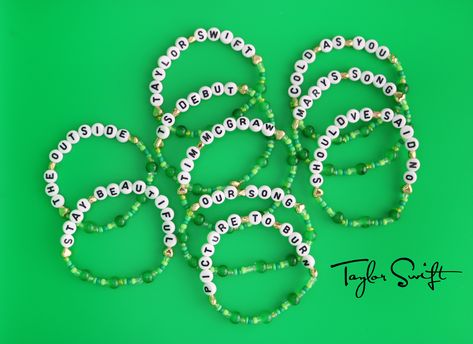 A spread of green beaded friendship bracelets with gold accents, made with stretchy string, glass beads, seed beads, and the names of the songs on Taylor Swift's Debut Album, written in alphabet beads. Taylor Swift Debut Album Bracelet, Debut Eras Tour Bracelet, Eras Tour Friendship Bracelets Debut, Green Taylor Swift Bracelet, Debut Bracelet Ideas, Taylor Swift Debut Bracelet Ideas, Debut Friendship Bracelet Ideas, Taylor Swift Debut Friendship Bracelet, Debut Taylor Swift Bracelet