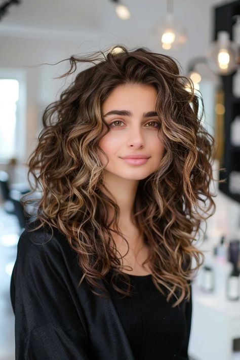 Woman with long, curly brown hair and a soft smile standing in a modern salon. Classy Haircolors, Babylights For Dark Brown Hair, Balayage Front View, Dark Caramel Balayage Brunettes, Brown Curly Hair With Highlights Caramel, Dark Curly Hair Balayage, Dark Brown Curly Hair Balayage, Caramel Highlights On Dark Brown Hair, Curly Brown Hair With Blonde Highlights