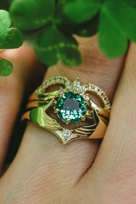 A close-up image shows the three-piece Claddagh ring set from Staghead Designs on a woman's ring finger. This 14K yellow gold set features a round cut lab-created emerald solitaire engagement ring, a crown-shaped tracer with diamond accents, & a tracer with two embracing hands & a pear cut diamond. Traditional Irish Wedding Rings, Irish Claddagh Engagement Ring, Three Ring Set, Celtic Engagment Rings, Engagement Rings Claddagh, Three Part Wedding Ring, Men’s Emerald Engagement Rings, Irish Wedding Rings Claddagh, Claddagh Wedding Ring Set